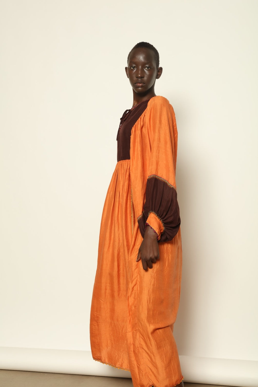 Model is wearing the Serafina Cognac Silk Dress by Supernaturae x HONS in warm cognac-toned handwoven silk, featuring a flowing kaftan-inspired silhouette and soft natural drape. Made with traditional artisan techniques and natural dyeing, this sustainable luxury dress has subtle tonal variations and adjustable side ties for a flexible fit, part of the House of Woven Stories selection.