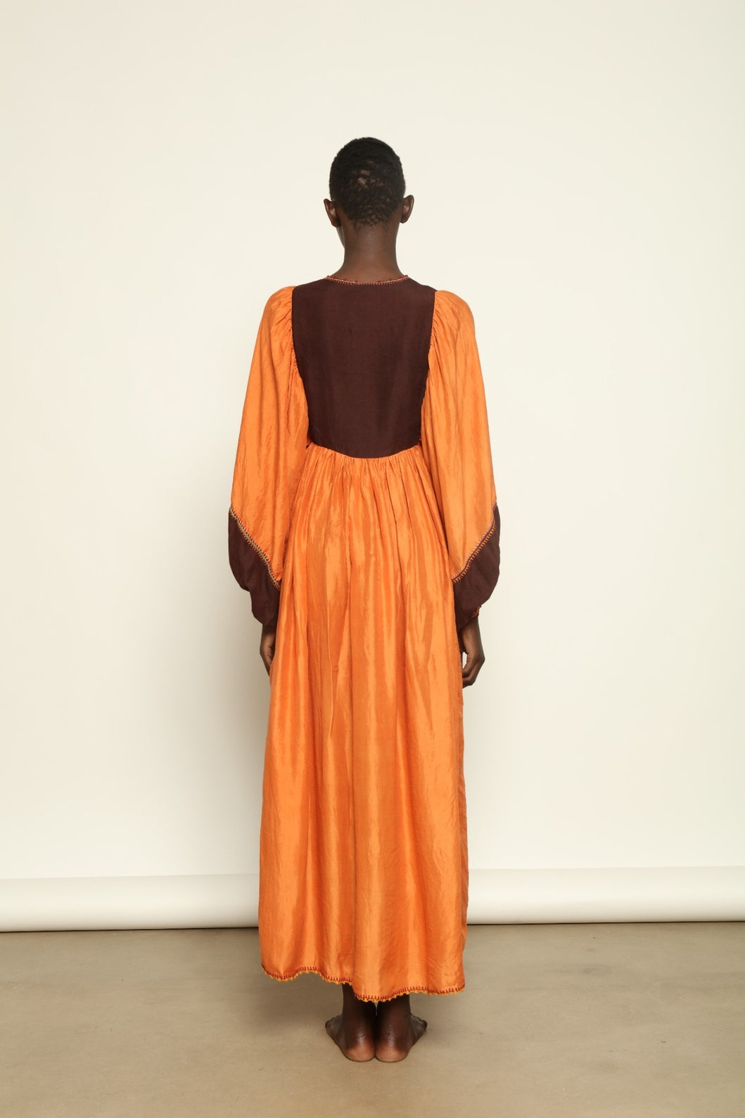 Model is wearing the Serafina Cognac Silk Dress by Supernaturae x HONS in warm cognac-toned handwoven silk, featuring a flowing kaftan-inspired silhouette and soft natural drape. Made with traditional artisan techniques and natural dyeing, this sustainable luxury dress has subtle tonal variations and adjustable side ties for a flexible fit, part of the House of Woven Stories selection.
