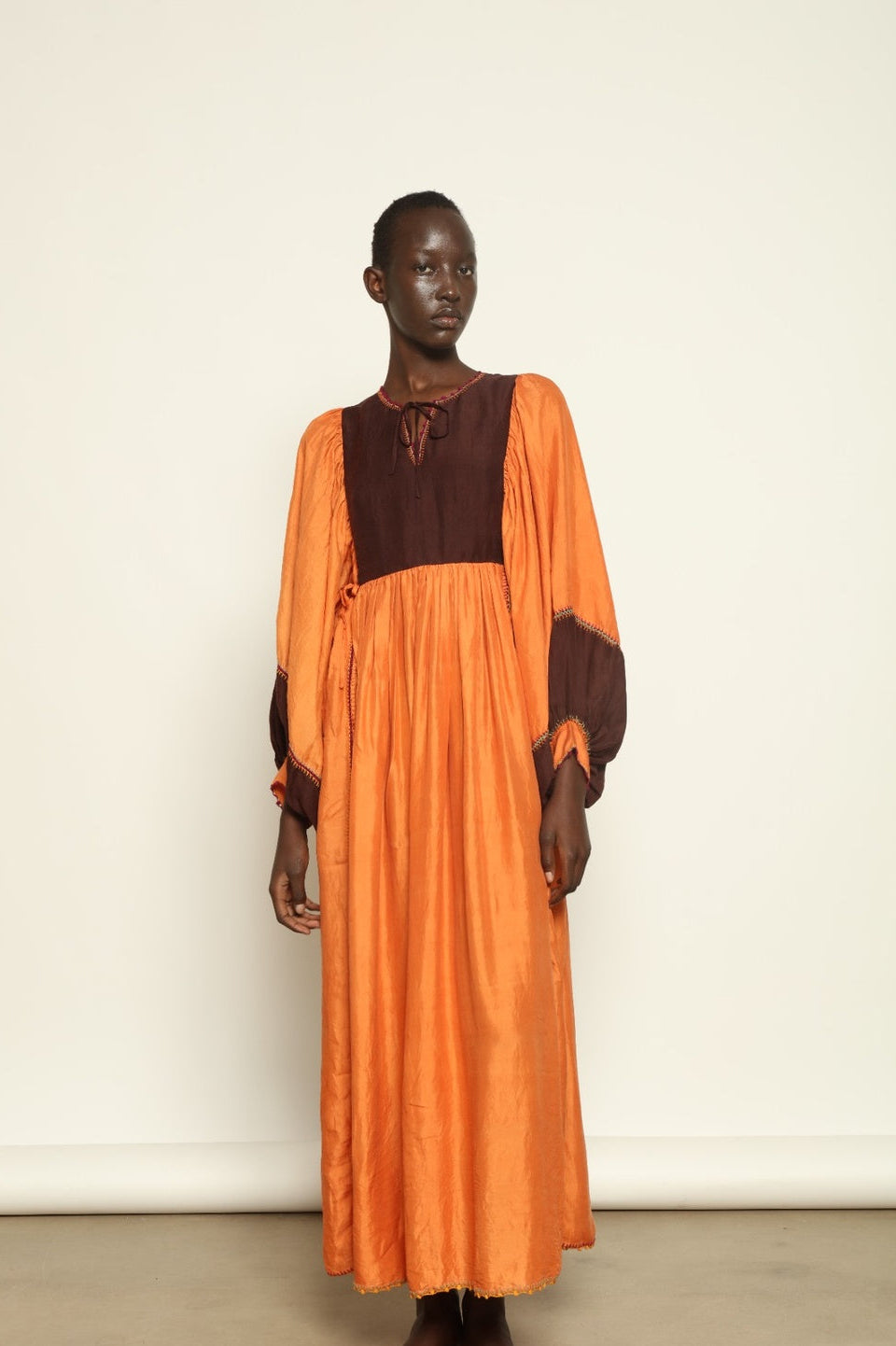 Model is wearing the Serafina Cognac Silk Dress by Supernaturae x HONS in warm cognac-toned handwoven silk, featuring a flowing kaftan-inspired silhouette and soft natural drape. Made with traditional artisan techniques and natural dyeing, this sustainable luxury dress has subtle tonal variations and adjustable side ties for a flexible fit, part of the House of Woven Stories selection.