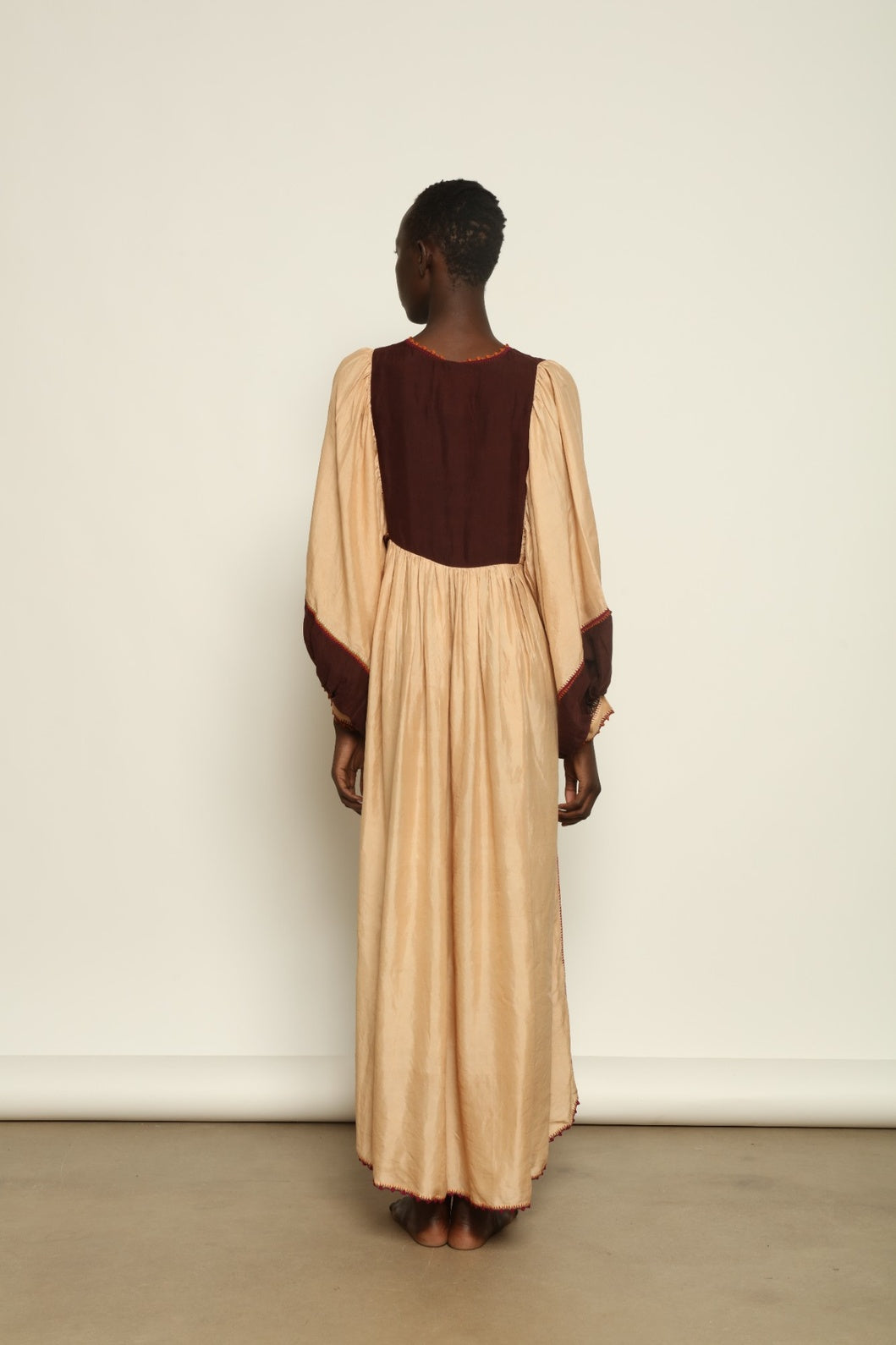 Model is wearing the Serafina Cream and Brown Silk Dress by Supernaturae x HONS, crafted in ethereal silk chiffon with cream and earthy brown botanical dyes. Featuring a 1970s-inspired flowing silhouette and delicate hand-crochet details, this sustainable slow luxury dress shows natural organic pattern variations created by plant-based pigments. Made in collaboration with artisan communities in Mexico, part of the House of Woven Stories selection