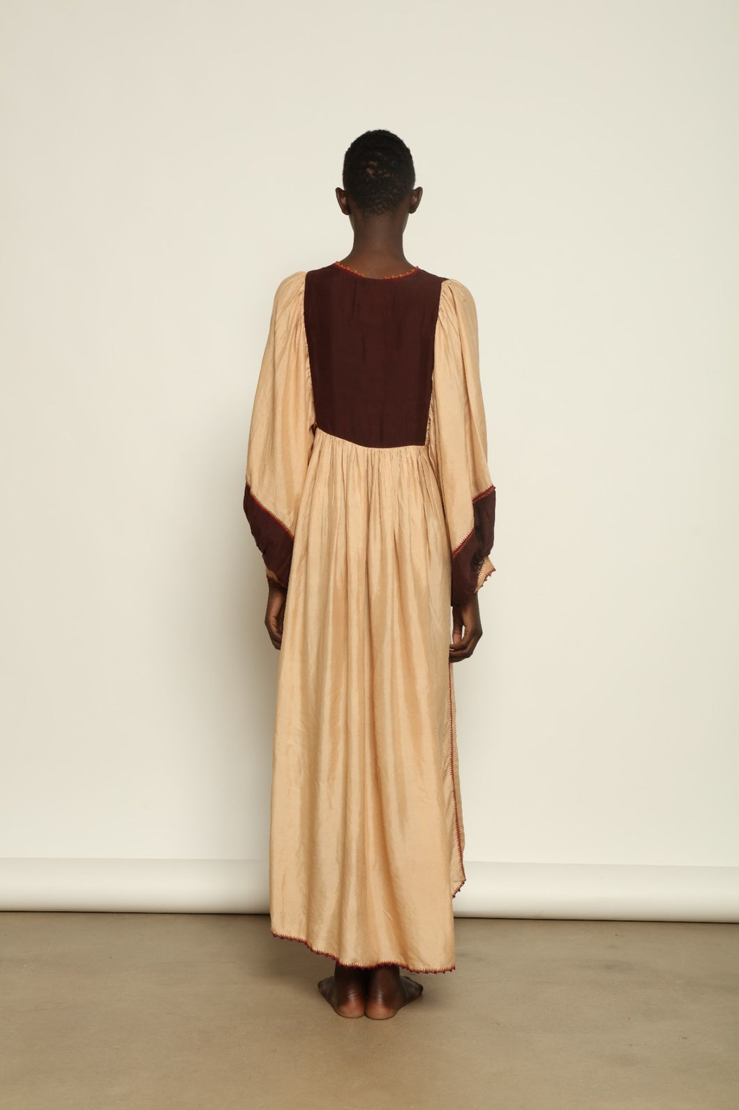 Model is wearing the Serafina Cream and Brown Silk Dress by Supernaturae x HONS, crafted in ethereal silk chiffon with cream and earthy brown botanical dyes. Featuring a 1970s-inspired flowing silhouette and delicate hand-crochet details, this sustainable slow luxury dress shows natural organic pattern variations created by plant-based pigments. Made in collaboration with artisan communities in Mexico, part of the House of Woven Stories selection