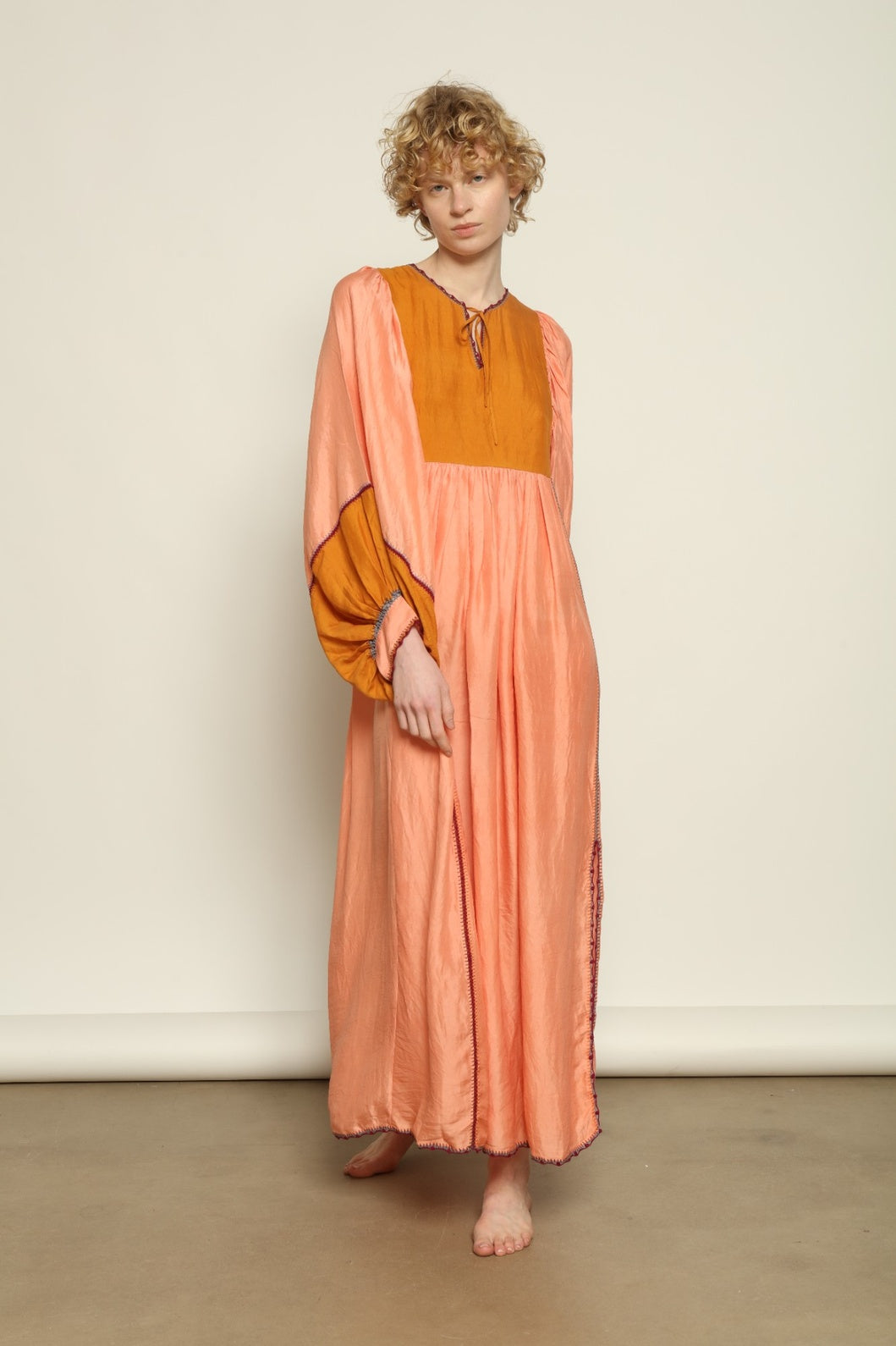 Model is wearing the Serafina Rose and Yellow Silk Dress by Supernaturae x HONS in pure handwoven silk, featuring a flowing kaftan-inspired silhouette with soft rose and golden yellow botanical-dyed tones. This sustainable artisan-made dress shows natural color variations unique to each piece, with lightweight breathable fabric and adjustable side ties, part of the House of Nomad Stories selection.