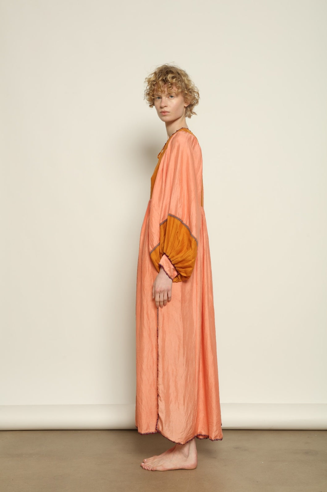 Model is wearing the Serafina Rose and Yellow Silk Dress by Supernaturae x HONS in pure handwoven silk, featuring a flowing kaftan-inspired silhouette with soft rose and golden yellow botanical-dyed tones. This sustainable artisan-made dress shows natural color variations unique to each piece, with lightweight breathable fabric and adjustable side ties, part of the House of Nomad Stories selection.