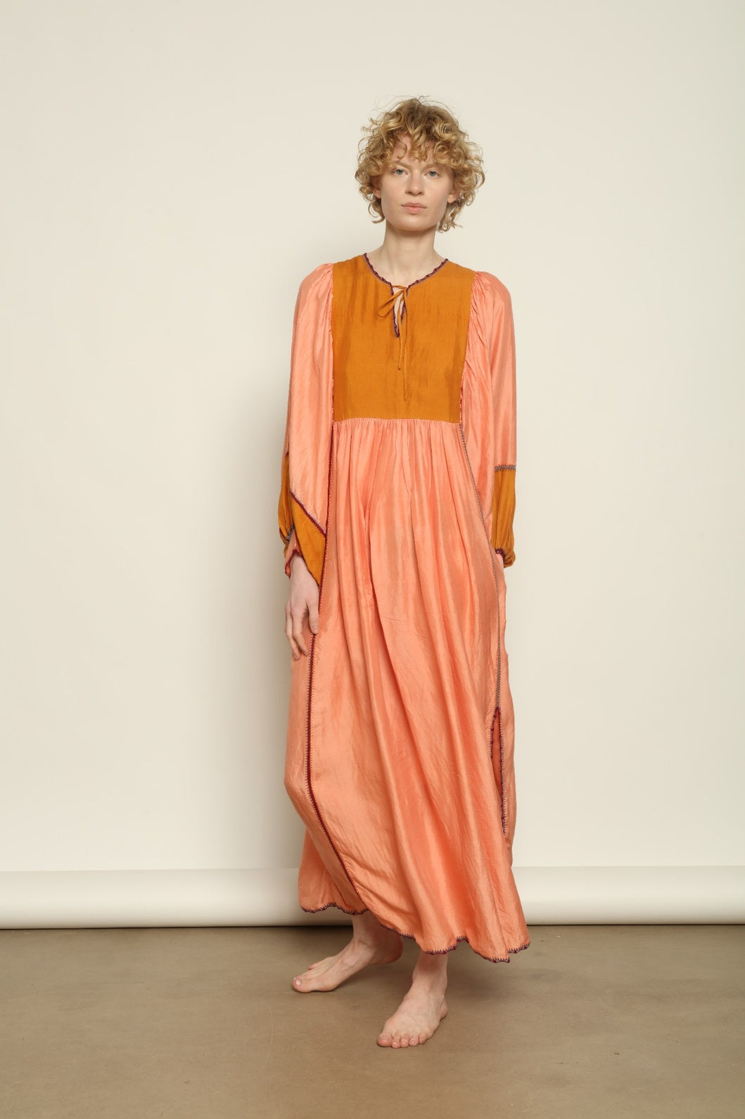Model is wearing the Serafina Rose and Yellow Silk Dress by Supernaturae x HONS in pure handwoven silk, featuring a flowing kaftan-inspired silhouette with soft rose and golden yellow botanical-dyed tones. This sustainable artisan-made dress shows natural color variations unique to each piece, with lightweight breathable fabric and adjustable side ties, part of the House of Nomad Stories selection.