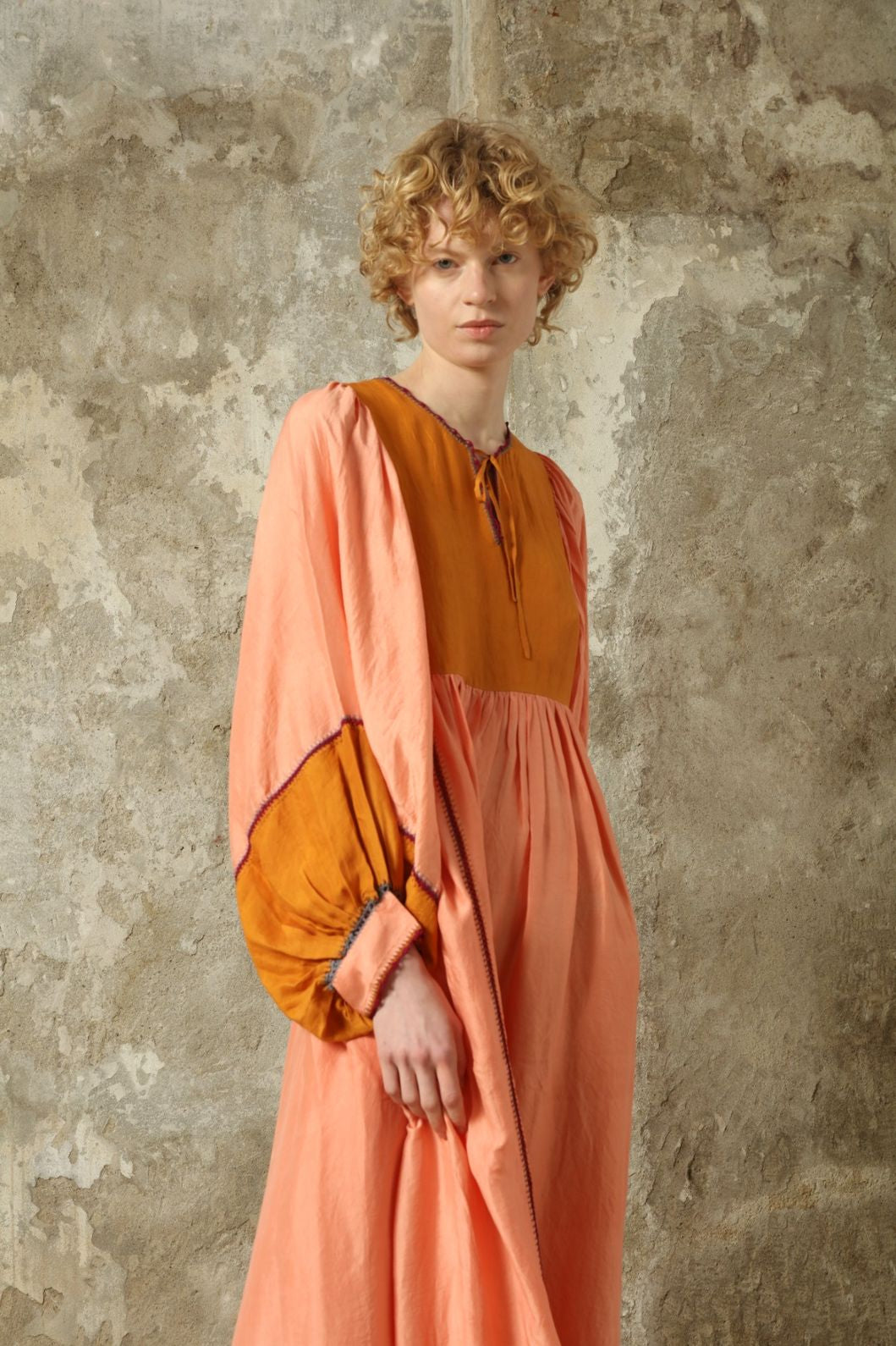 Model is wearing the Serafina Rose and Yellow Silk Dress by Supernaturae x HONS in pure handwoven silk, featuring a flowing kaftan-inspired silhouette with soft rose and golden yellow botanical-dyed tones. This sustainable artisan-made dress shows natural color variations unique to each piece, with lightweight breathable fabric and adjustable side ties, part of the House of Nomad Stories selection.