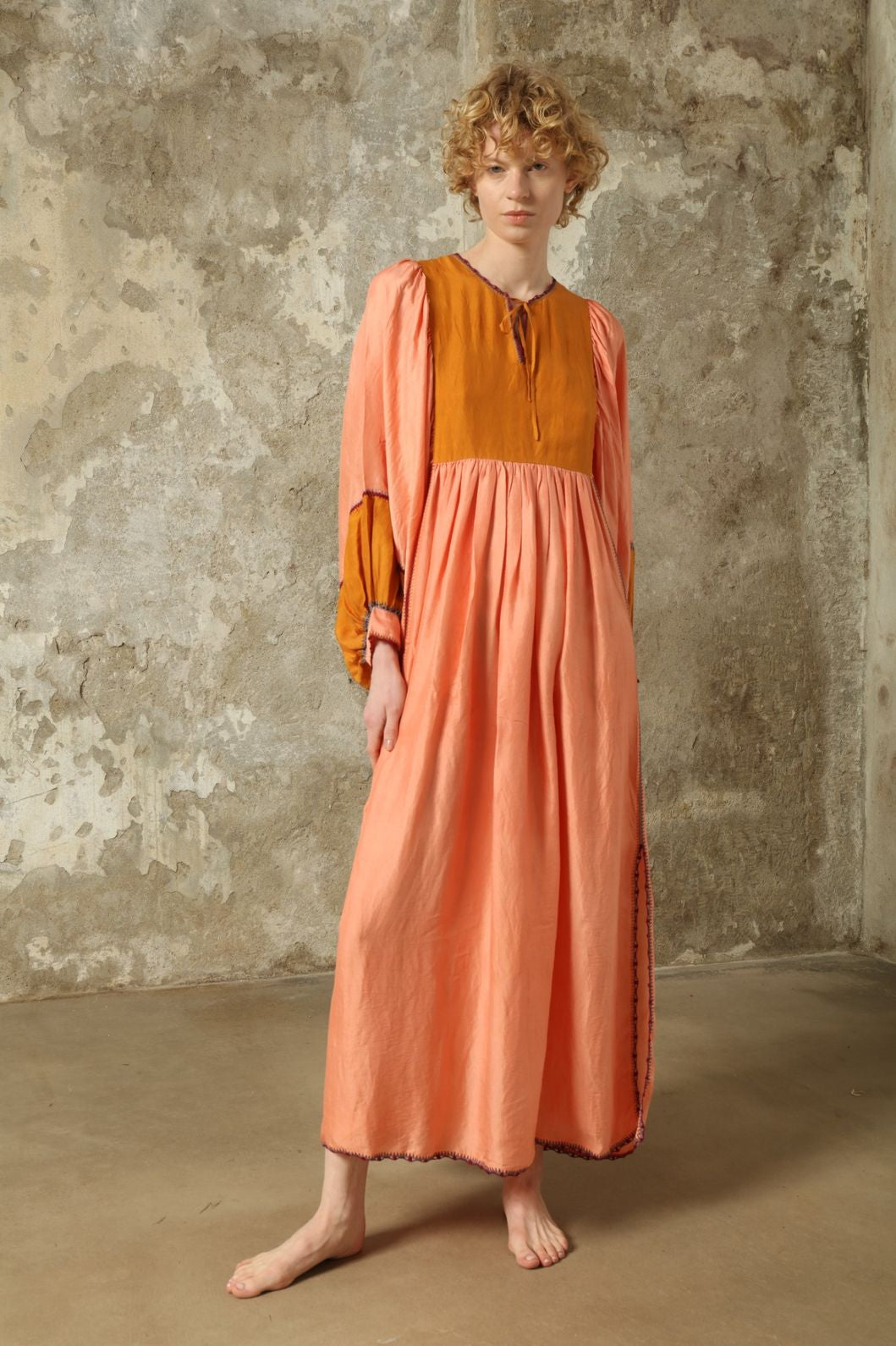 Model is wearing the Serafina Rose and Yellow Silk Dress by Supernaturae x HONS in pure handwoven silk, featuring a flowing kaftan-inspired silhouette with soft rose and golden yellow botanical-dyed tones. This sustainable artisan-made dress shows natural color variations unique to each piece, with lightweight breathable fabric and adjustable side ties, part of the House of Nomad Stories selection.