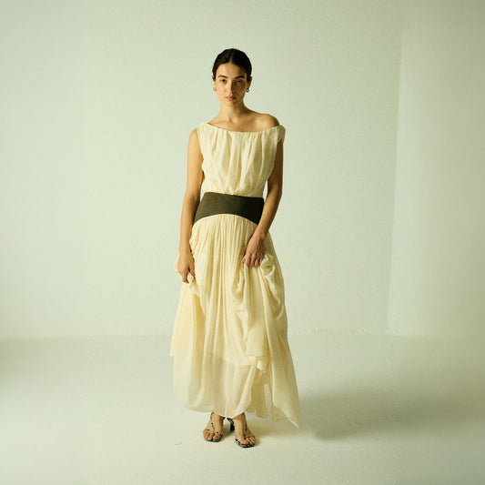 Model wearing a cream Siena Maxi Dress by Abadia from the House of Nomad Stories collection, featuring an off-shoulder draped bodice, sculpted green waistband, and a flowing elegant maxi silhouette.