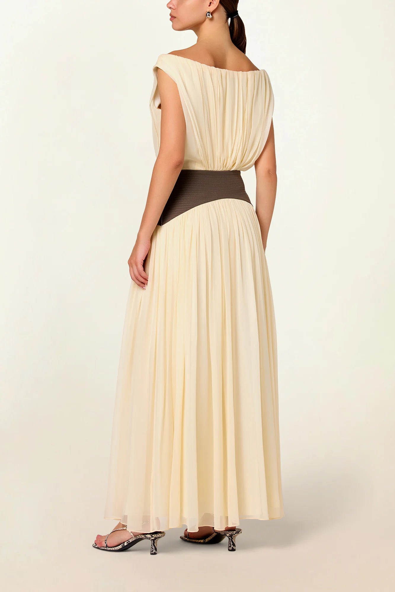 Model wearing a cream Siena Maxi Dress by Abadia from the House of Nomad Stories collection, featuring an off-shoulder draped bodice, sculpted green waistband, and a flowing elegant maxi silhouette.