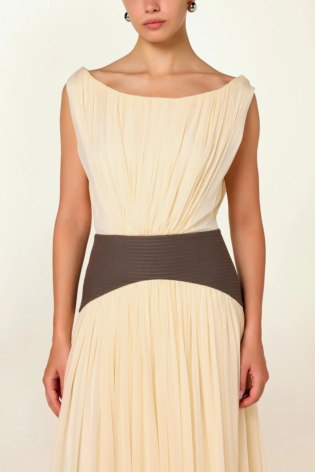 Model wearing a cream Siena Maxi Dress by Abadia from the House of Nomad Stories collection, featuring an off-shoulder draped bodice, sculpted green waistband, and a flowing elegant maxi silhouette.