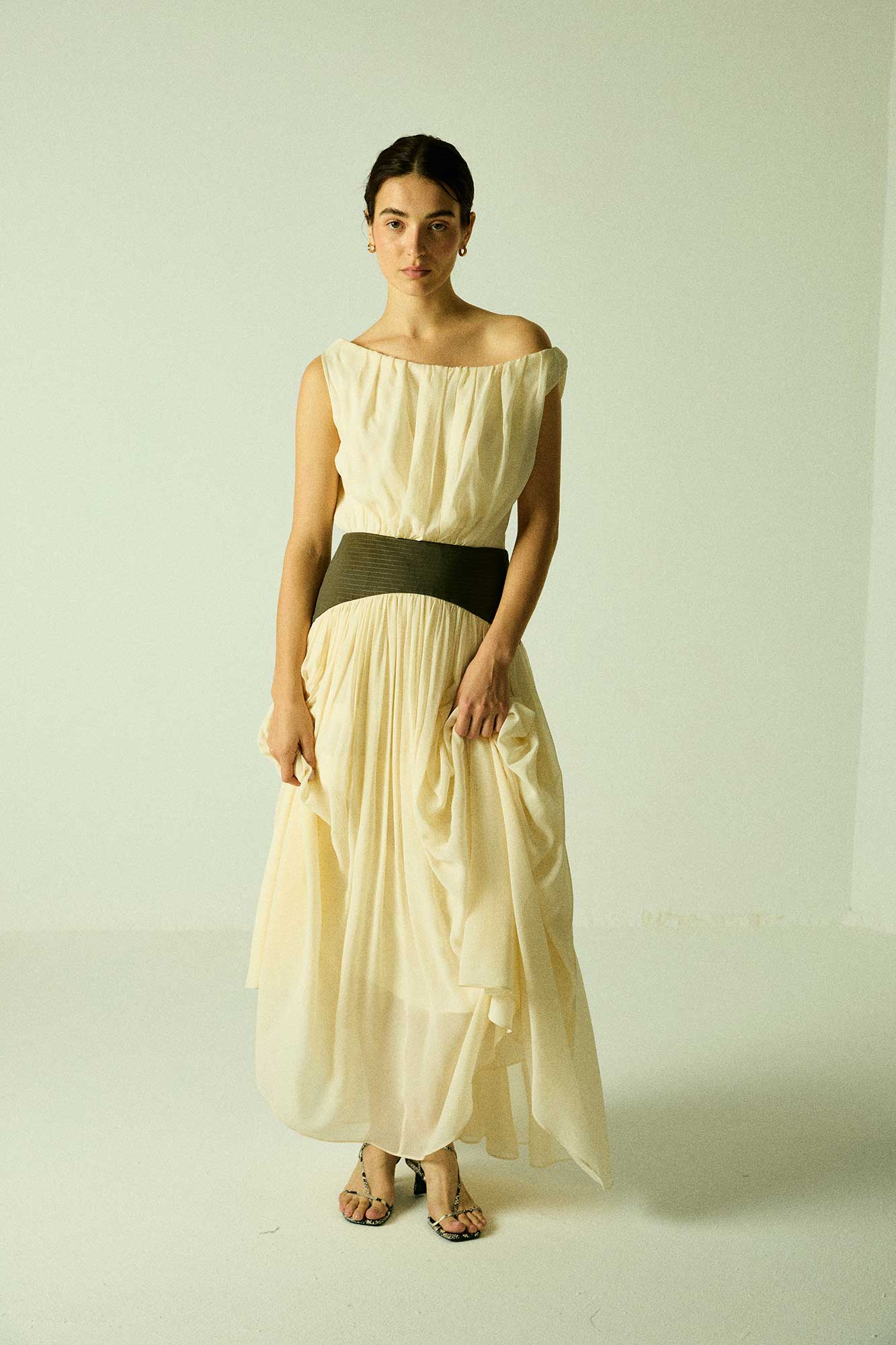 Model wearing a cream Siena Maxi Dress by Abadia from the House of Nomad Stories collection, featuring an off-shoulder draped bodice, sculpted green waistband, and a flowing elegant maxi silhouette.
