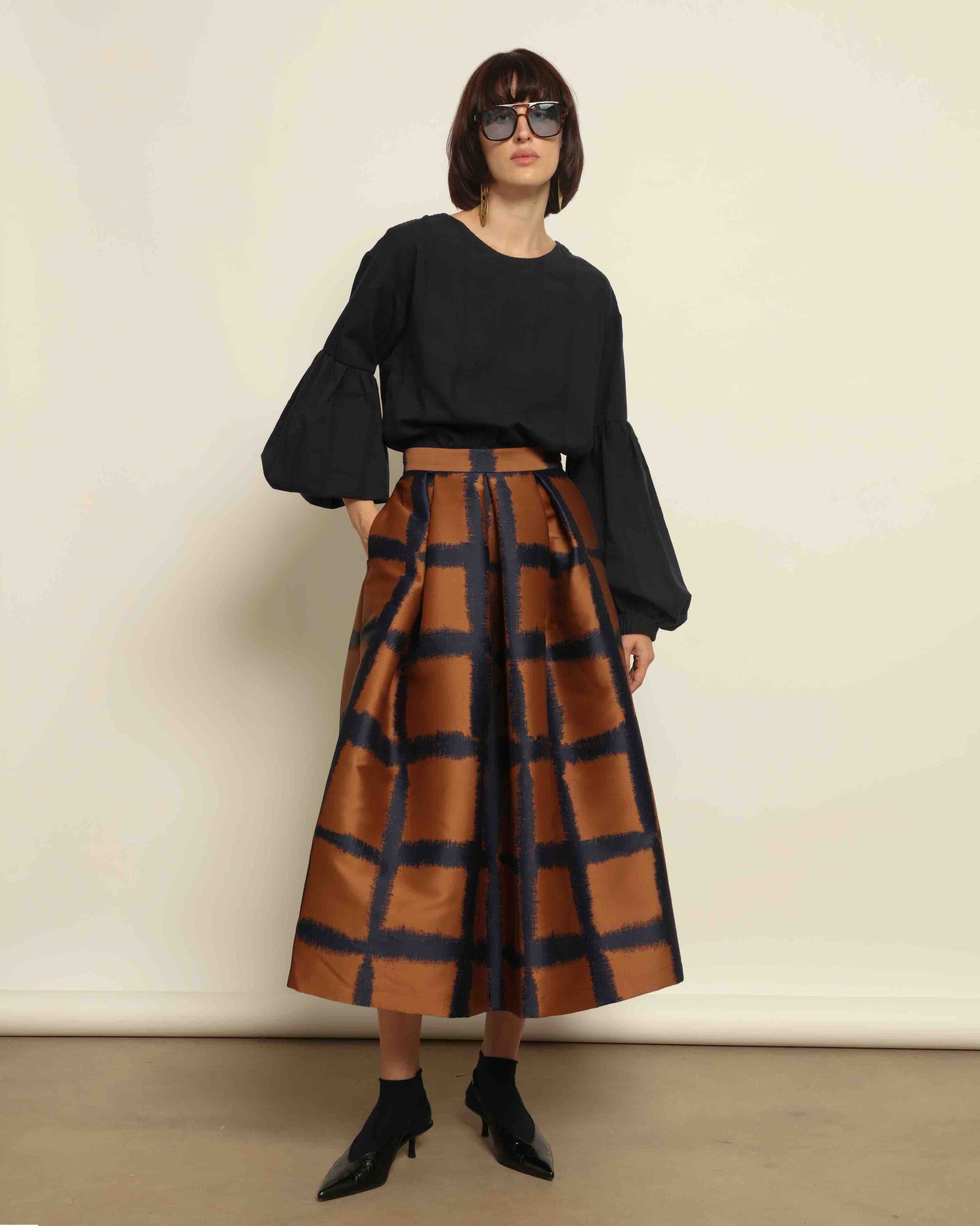 Model is wearing the Square Orange and Blue Skirt by Lunatica Milano. Pleated skirt in bronze and deep blue with striking geometric design. Feminine, elegant, and handcrafted in Italy. Part of House of Woven Stories selection.